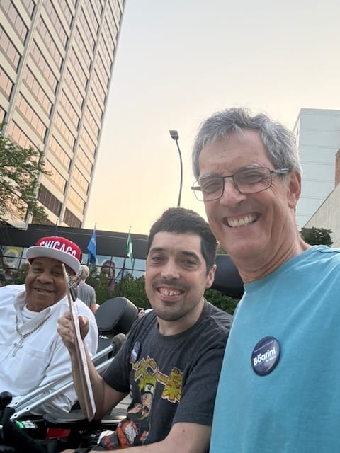 Jeff Boarini, wearing a campaign button, smiling in a selfie with his son Ted and a man in a Chicago cap on a downtown sidewalk at sunset, out collecting petition signatures